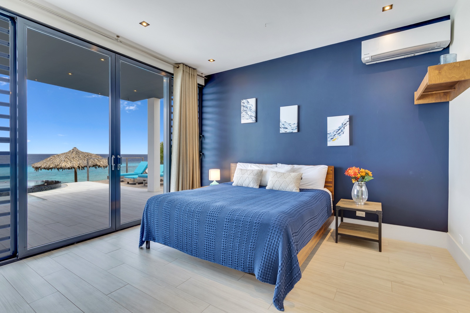 Luxury Bonaire villa master suite with a blue accent wall, king bed, and floor-to-ceiling sliding glass doors opening to a private terrace with panoramic ocean views.